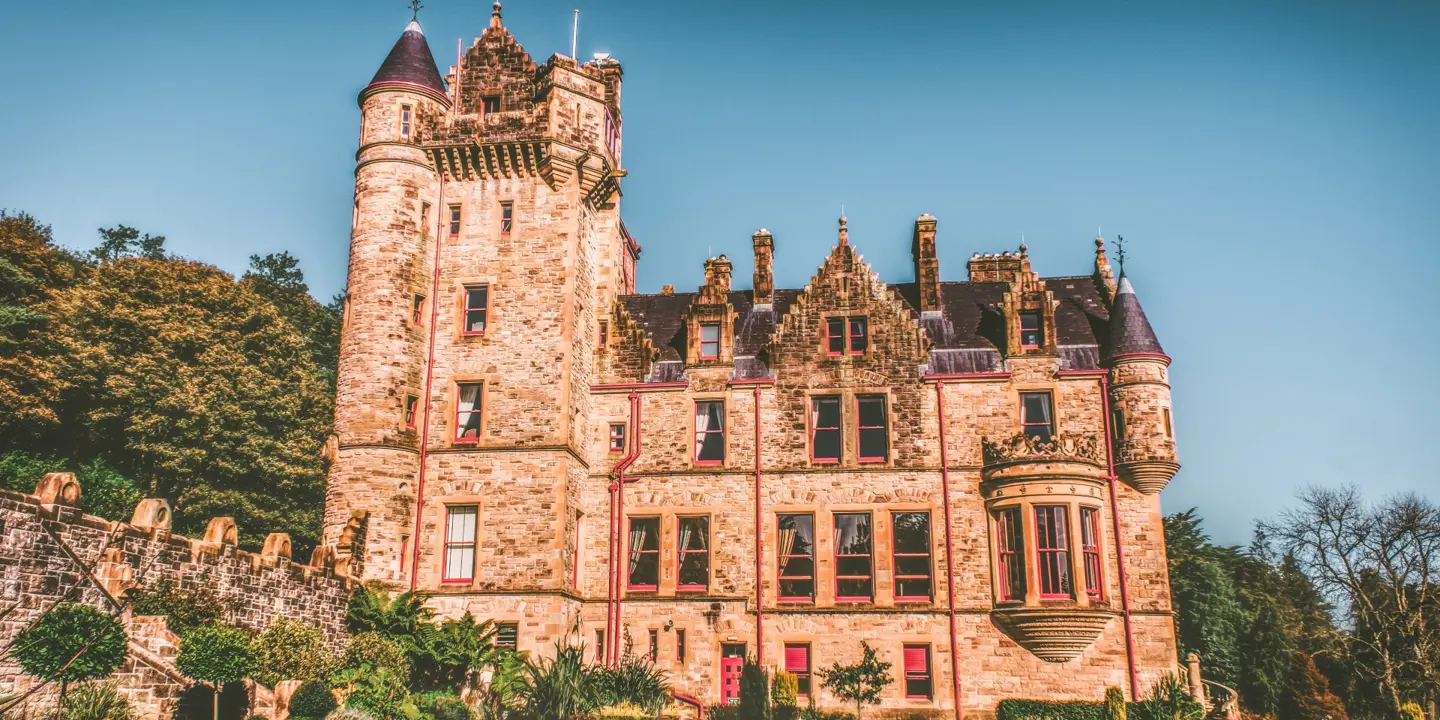 A view of Belfast castle in warm sunlight highlighting the distinctive brick colours and the red guttering on the building. Well manicured lawns are visible as well as trees in full bloom.