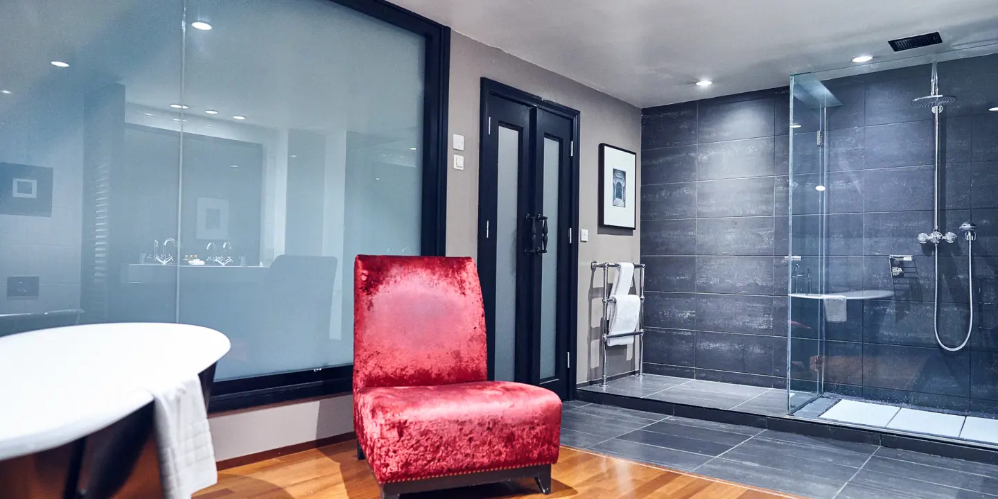 Bathroom featuring walk in shower, bathtub and red velvet chair.