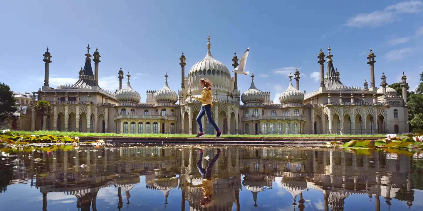 A girl in a yellow top and blue jeans runs across the pond in front of the Brighton Pavilion with a seagull in flight behind her. The image is reflected in the pond.