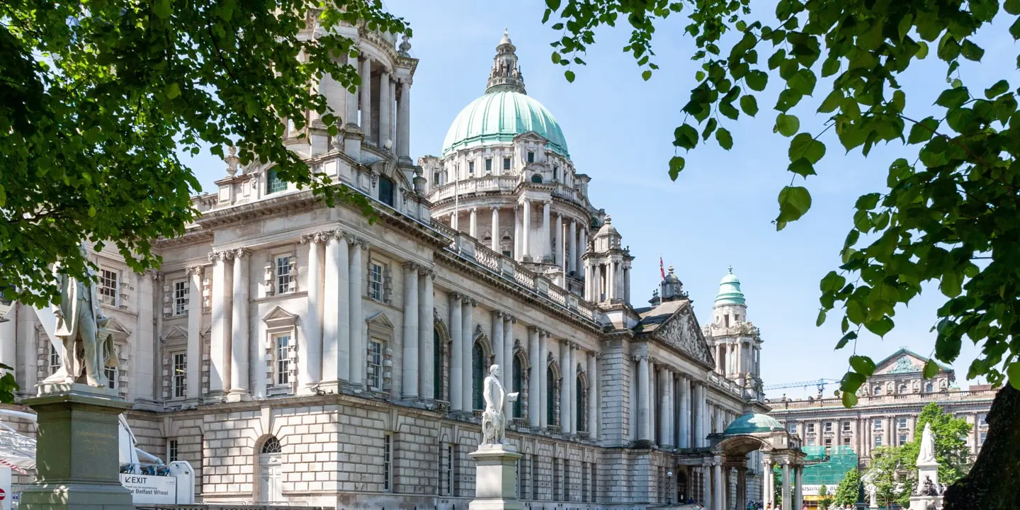A view of Belfast city hall with several statues visible in the lush green grounds, park benches line a well kept pathway. The architecture of the building and the turquoise colouring of the once copper roof are prominently visible.