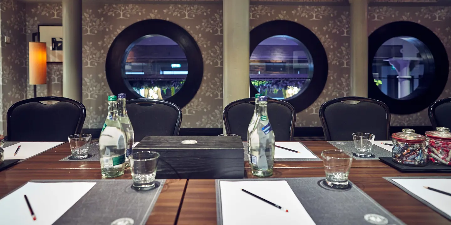 Luxurious meeting room showing a close up of a wooden table adorned with bottles of water and glasses, with 5 black chairs in focus, and 3 black framed porthole style windows in the background.