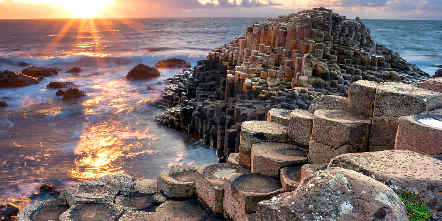 The giants causeway at sunset, the sun is just above the sea on the horizon and glints on the surf of the waves lapping at the rocks.