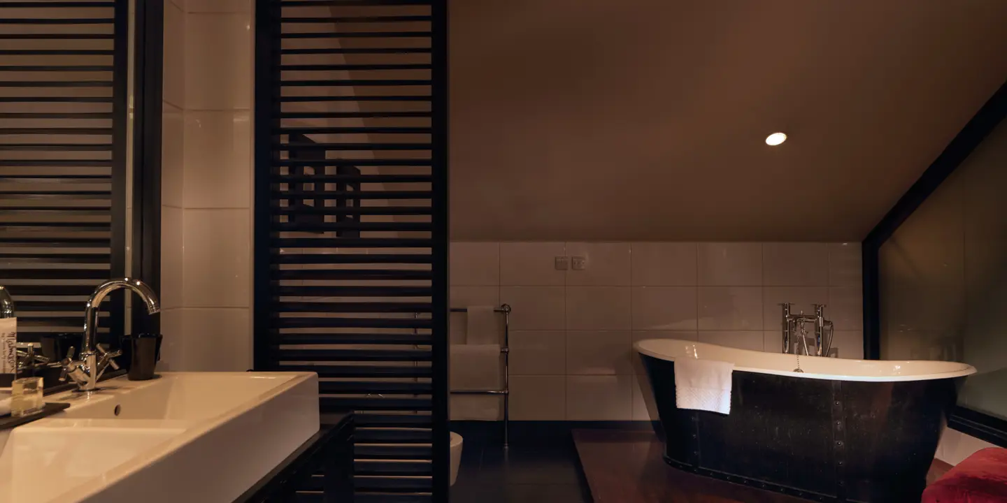 A darkly lit bathroom, with a free standing black bath tub and heated towel rail.