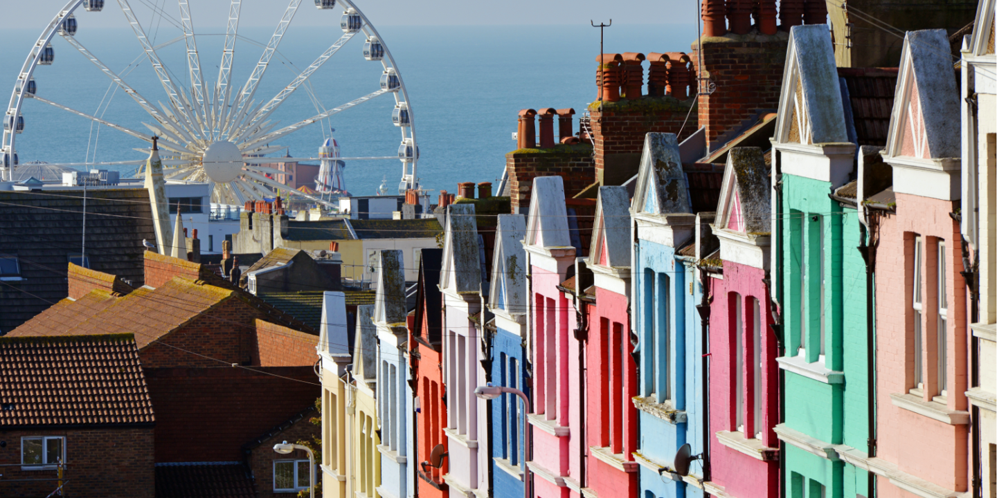 A row of vibrantly coloured terrace houses overlooking Brighton seafront. A ferris wheel is visible in the background along with the helter skelter on Brighton pier.