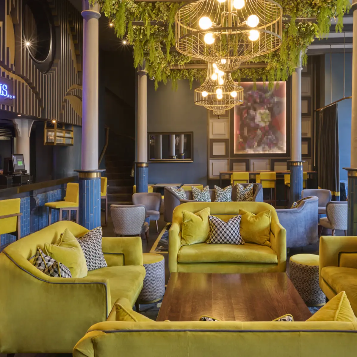 Yellow fabric sofas and bar stools prominently feature around a wooden table, with the bar in the background and a blue neon sign reading "happiness is" is displayed above the bar.